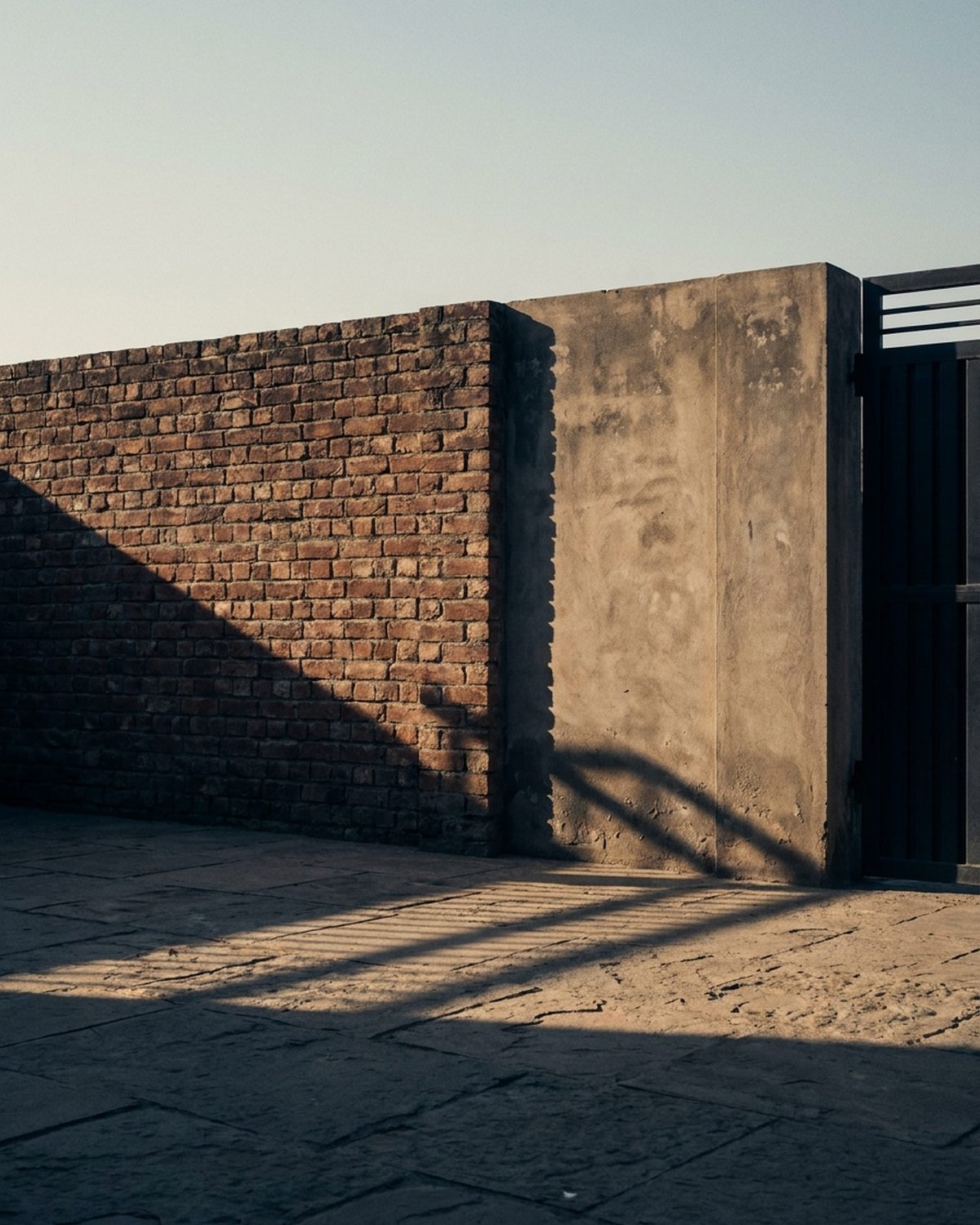 Brick boundary wall meeting a concrete gate pier and paved courtyard in late afternoon light